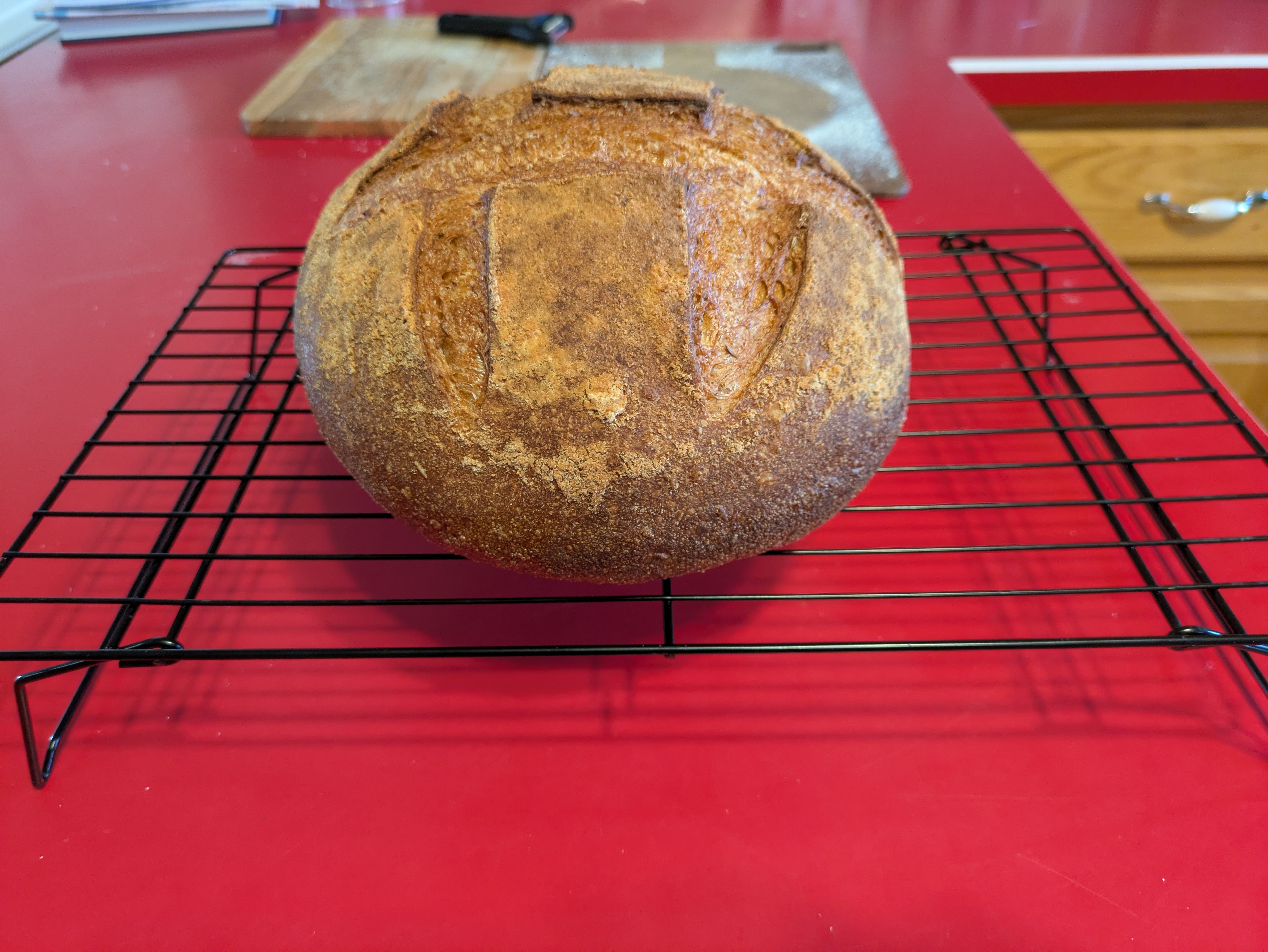 A loaf of bread in a boule shape on a black wire cooling rack. The bread is light to dark brown, with some score lines across the top of the boule.