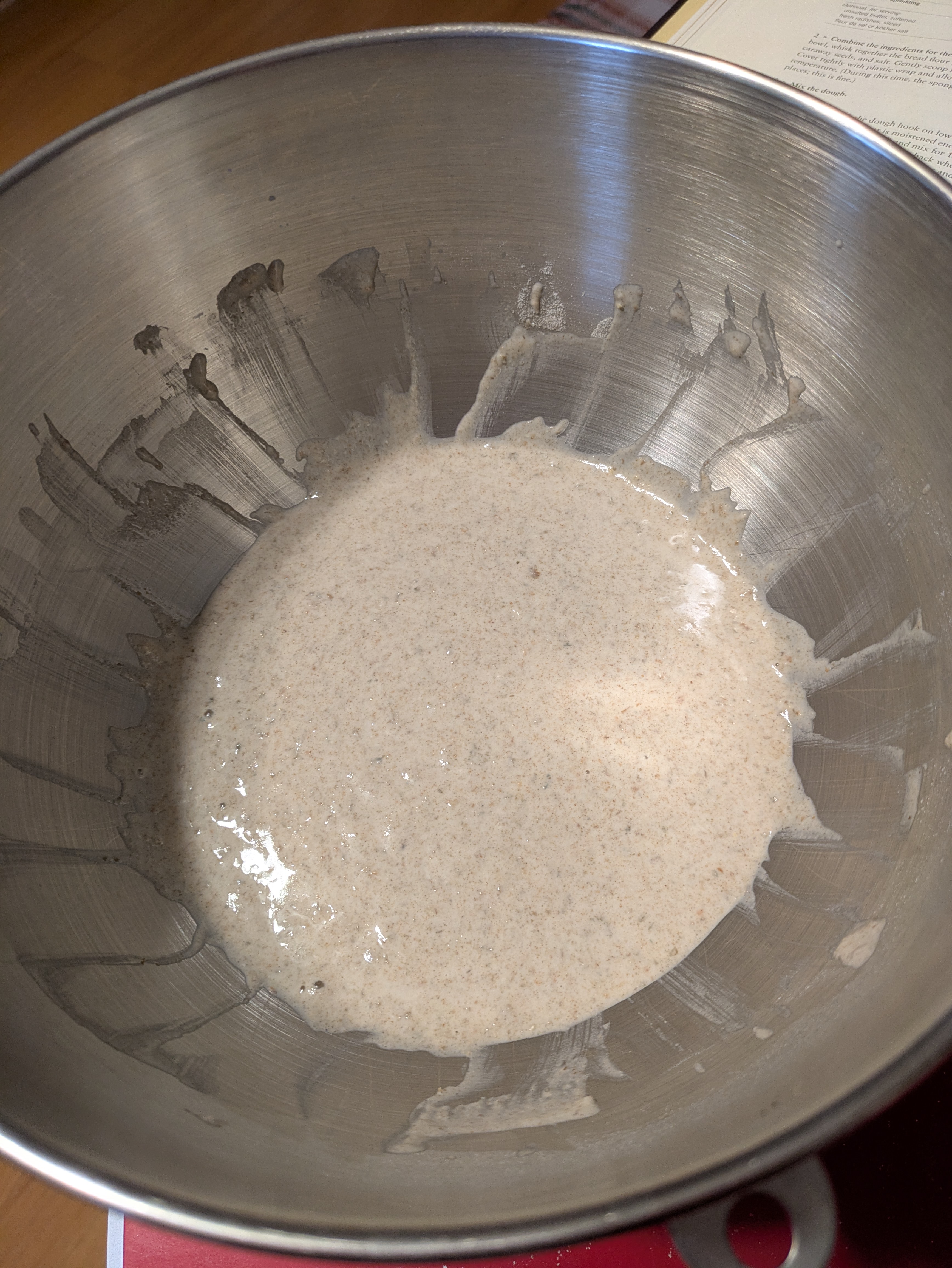 A photograph of a mixing bowl taken from above the bowl, looking down directly into its contents. In the bowl is a speckled, beige to light brown liquid batter - the sponge preferment - with a few air bubbles visible.