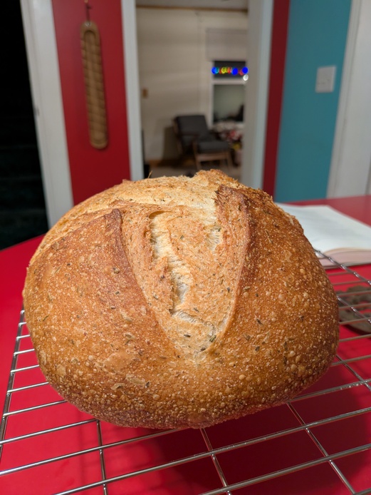 A round loaf (boule) of bread on a wire cooling rack, atop a red counter. The bread ranges from very light tan to golden brown. The crust is cracked a bit at the score line, which comes down the center of the loaf in a 'V' shape.