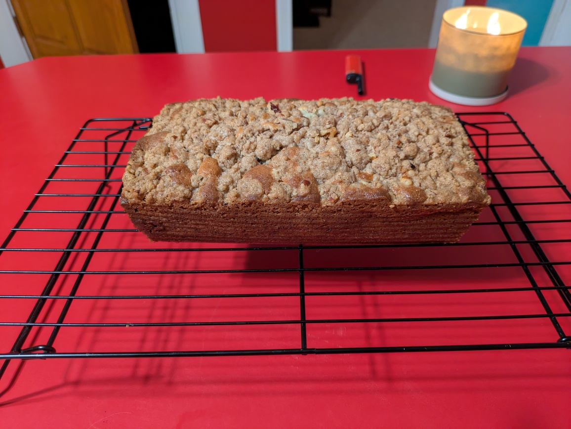 A short loaf of bread with a crumble topping on a black wire rack. The photograph is taken from above, at an angle. The rack is on a red counter.