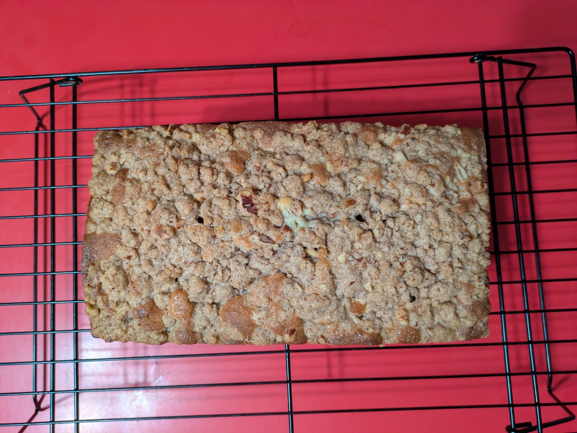 A photograph of the crumble topping on a loaf of bread, taken from directly atop the bread so that all you see is the crumble. The bread is on a black wire rack against a red counter background.