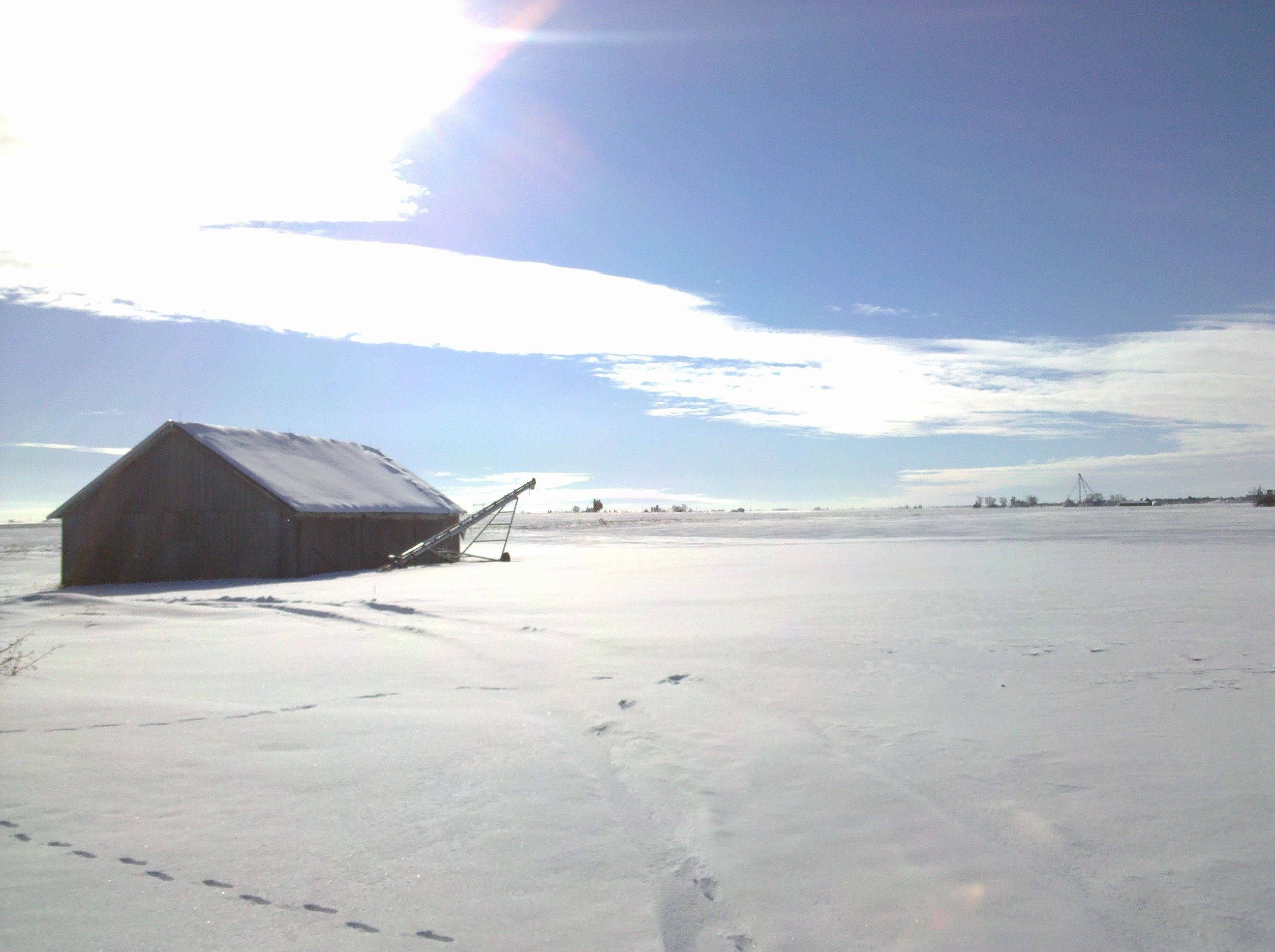 A snowy landscape. There is a farm building in the foreground but little else. The land is mostly empty but for snow. The sky is baby blue with a long swoop of bright white clouds arching from the top-left to the right of the frame.