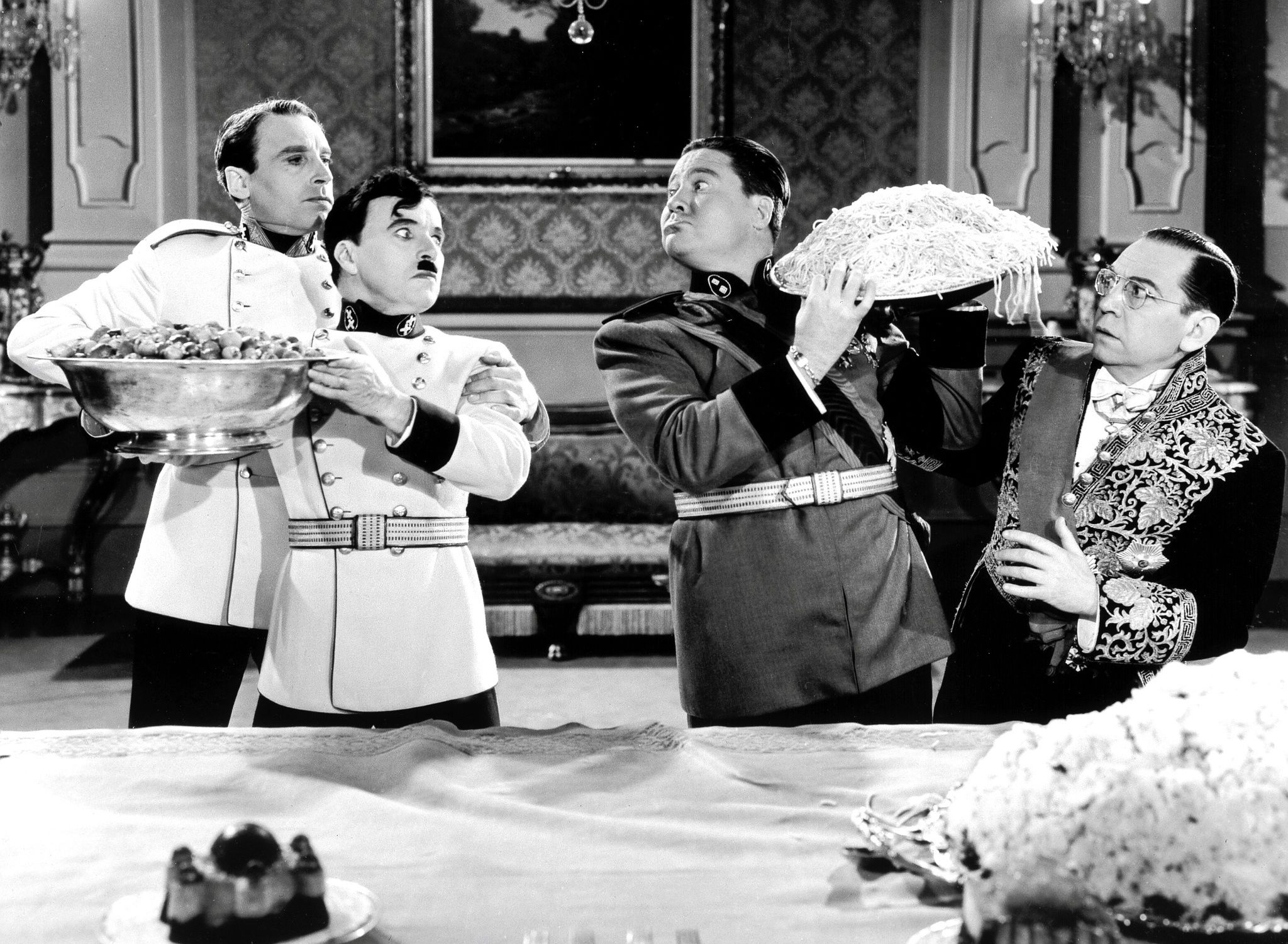 A still from the film 'The Great Dictator.' shows an ensemble of men in military uniform. From left to right: a man in a white military uniform holding a serving tray, a shorter man with a distinctive small mustache in a dark jacket, a heavyset man in military dress uniform holding what appears to be a large tray of pasta, and a man in ornate formal wear or costume on the right. The setting appears to be an elegant interior with ornate wallpaper and furnishings visible in the background. All are looking at each other with looks that range from surprise to frustration.