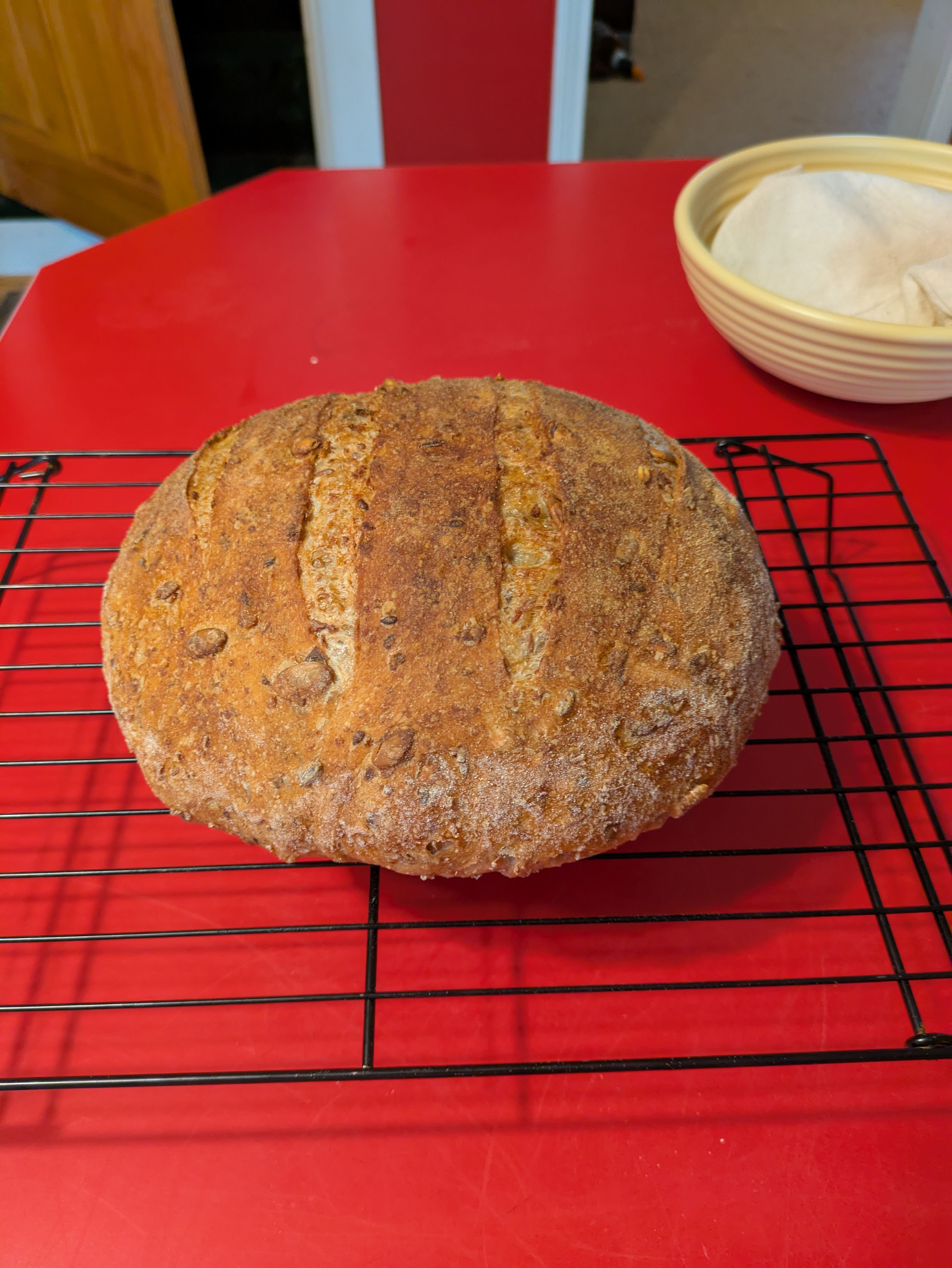 Small boule on a wire cooling rack. Some seeds are visible on the top of the boule. The crust is golden brown. The rack and bread sit upon a red countertop.