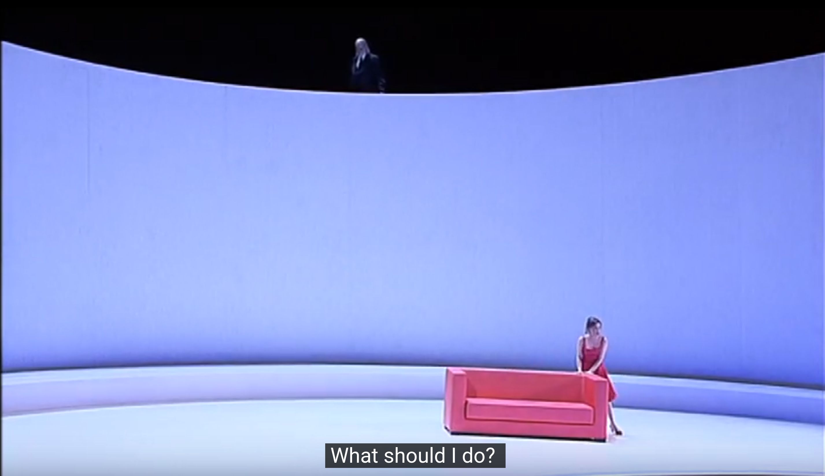A screenshot of a staged performance of La Traviata. The camera is at a fair distance from the stage, looking down. Violetta is in the center, in a red dress behind a red couch. At the very top of the frame, a man in a suit looks down at the scene. The caption reads: 'What should I do?'