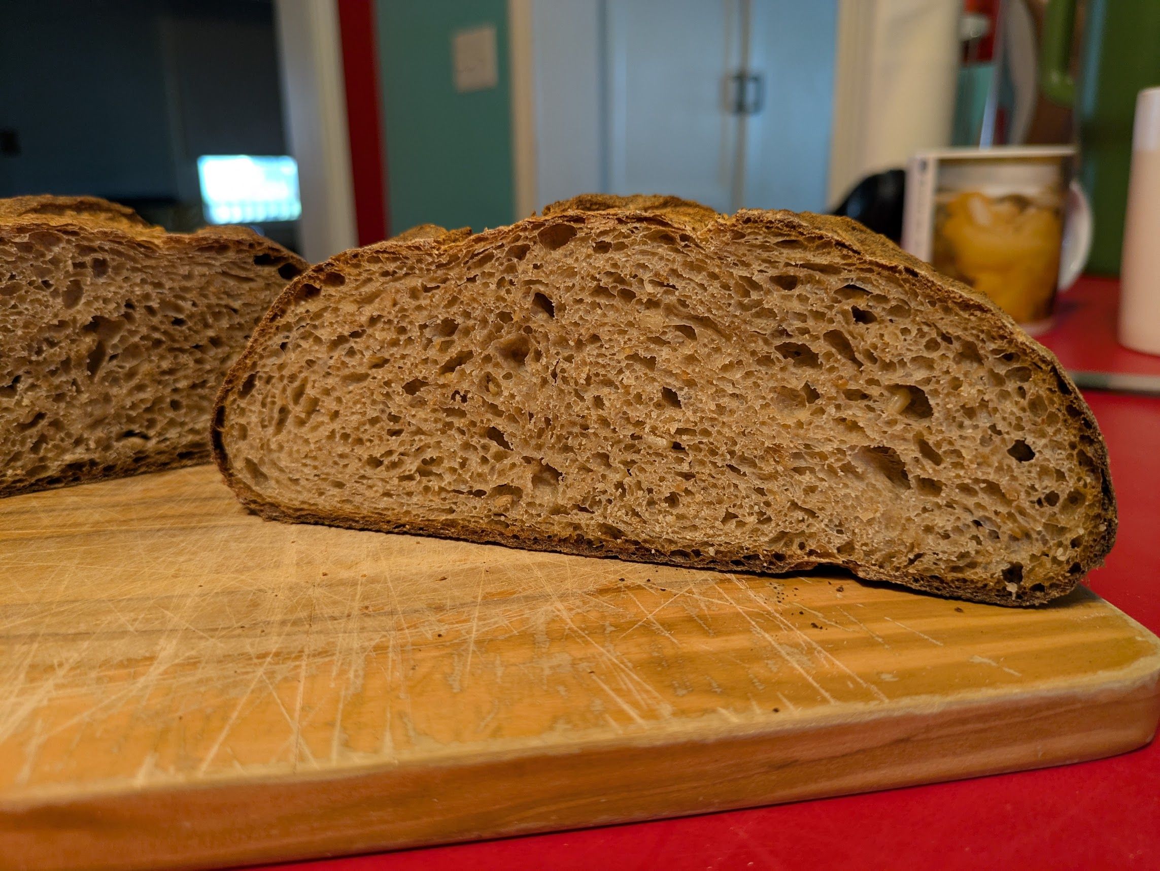 Half a loaf of bread, cut-side facing the camera, resting on a cutting board. The other half is similarly positioned and visible at the edge of the frame. The tan crumb of the bread is fairly open with a dark brown crust.