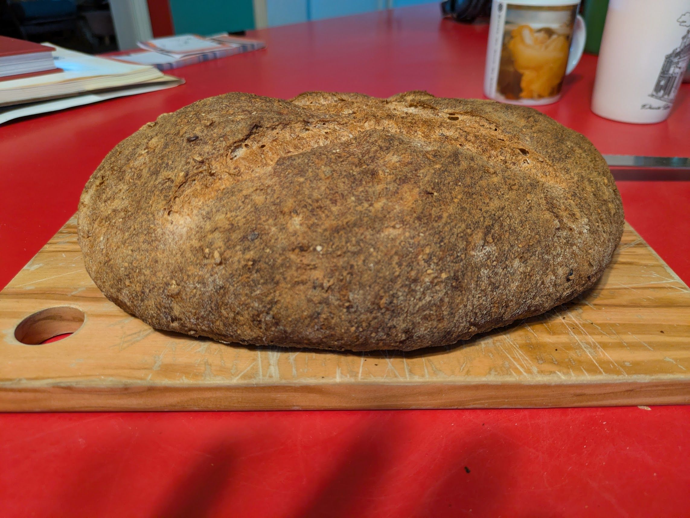 An oval-ish loaf of bread on a cutting board. The bread is dark tan to brown and the crust has a fairly rough exterior.