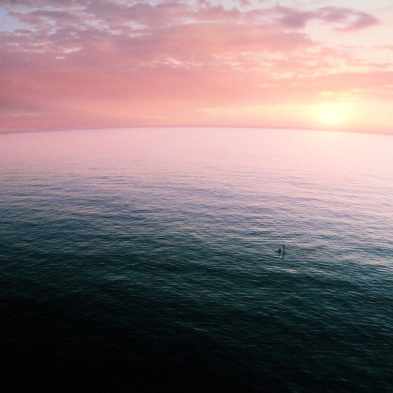 A photograph of a vast open ocean and sky. The ocean takes up the bottom 2/3 of the frame and shifts from deep dark in the bottom left, through blue, to a bright white-pink near the horizon. The hazy sky is cloudy with shades of mauve and lavender. A small speck can be made out as paddleboarders in the middle of the ocean's expanse.