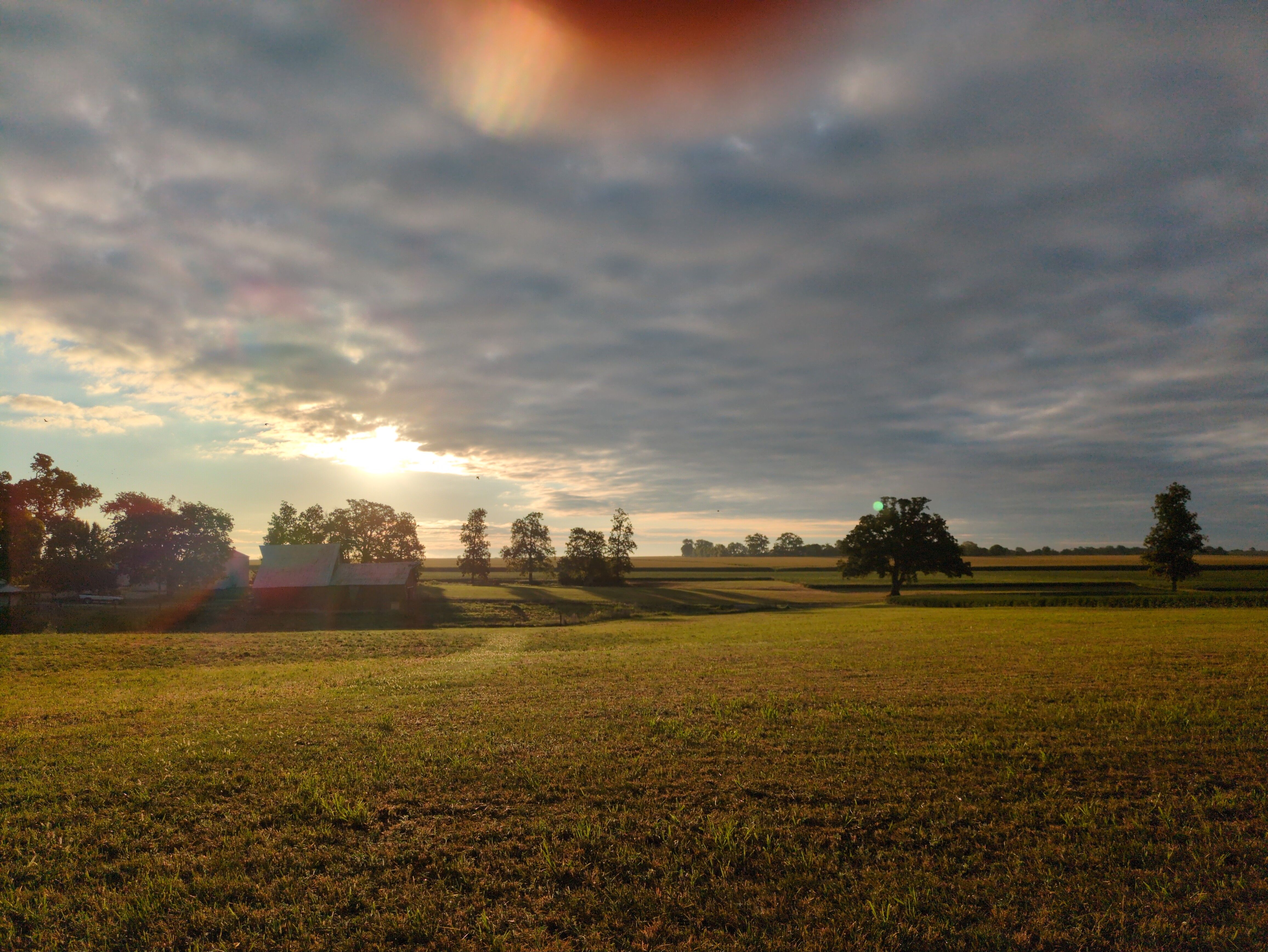 A landscape photo. A wide expanse of mowed field and grass, and a barn and trees in the distance. Much of the sky is blanketed in cloud cover. Toward the left of the frame, above the barn, the sun is shining against a small portion of clear sky. A treeline is visible further in the distance, and a bit of a fingertip has made its way into the top of the frame.