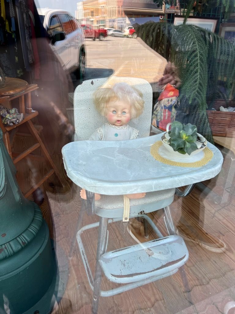 A vintage baby doll with light hair and blue eyes sits in a light blue metal high chair with a matching tray. The high chair appears to be from several decades ago. The scene is photographed through a display window, creating reflections that show cars and buildings in the background. The high chair is positioned on a wooden floor, and there are various other vintage items visible in the background including urniture and decorative objects.