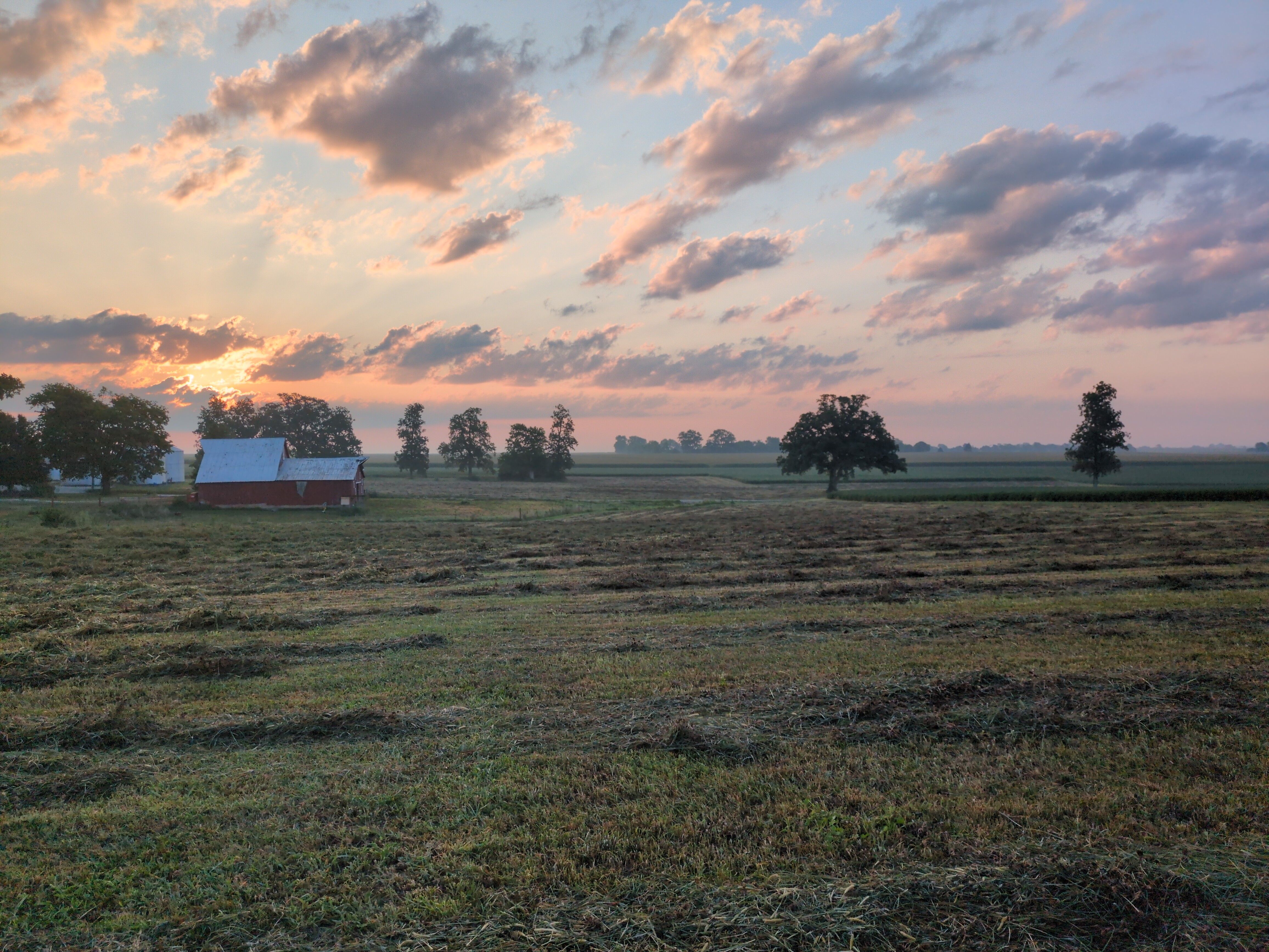 A landscape photo. A field, recently mowed down, and a barn and trees in the distance. The sky is dappled with clouds and, toward the left of the frame, the sun is shining through, turning that portion of the sky interesting colors of orange and pink. The silhouette of a treeline is visible further in the distance.