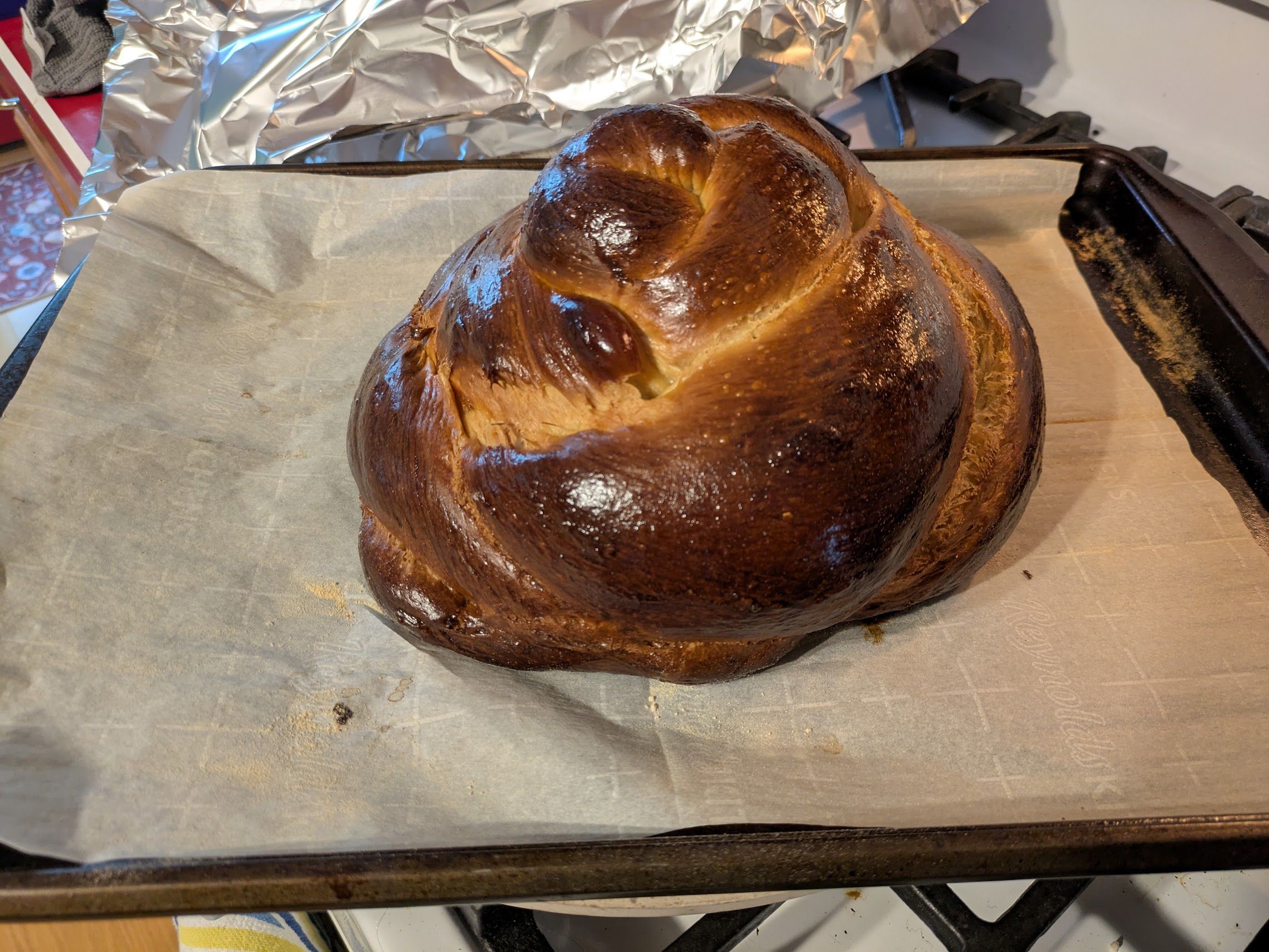 A very dark rounded strand loaf of challah, on a parchment-lined baking sheet.