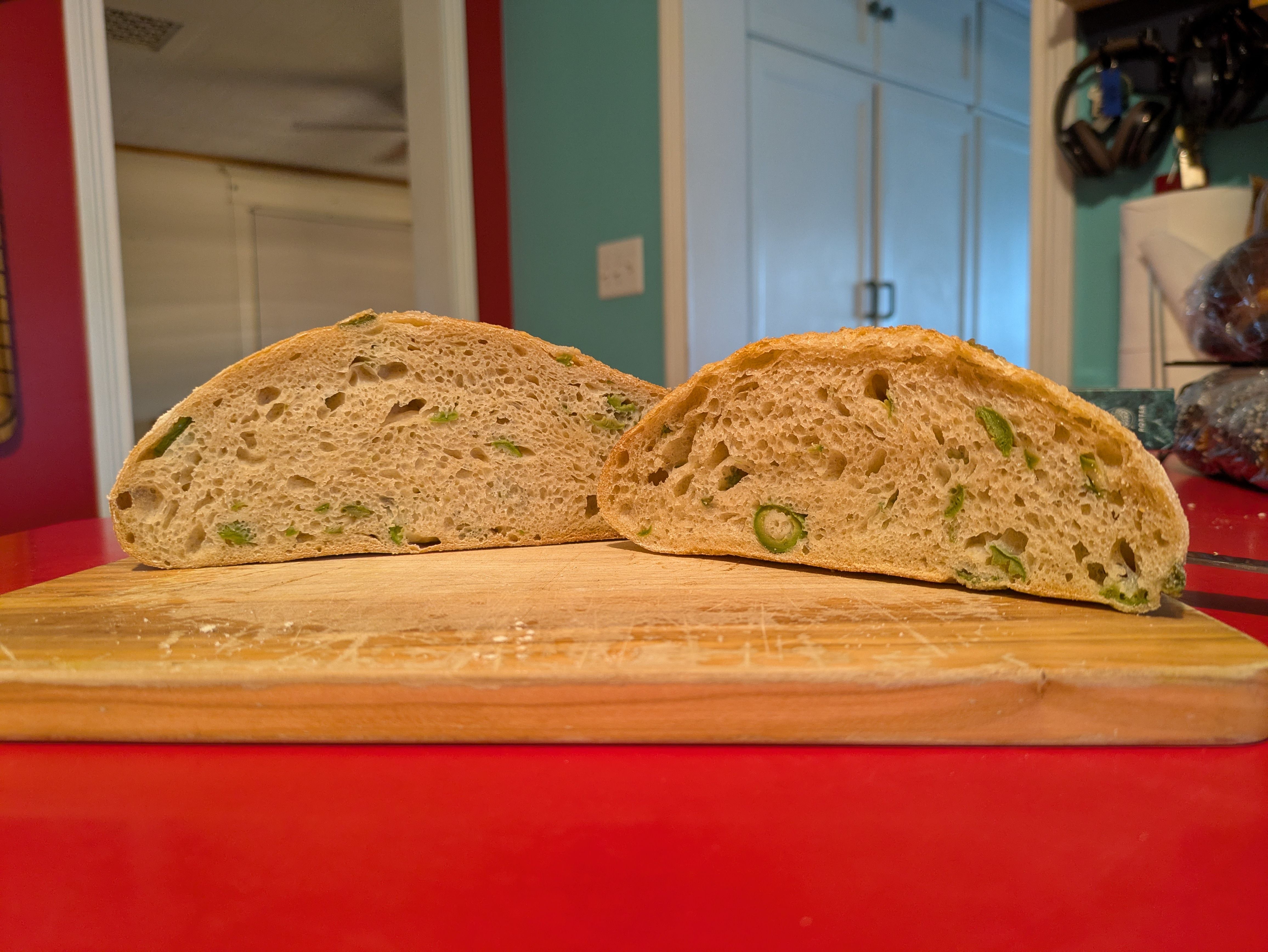 Two halves of a boule, interior (crumb) facing the camera. Bits of green olive can be seen across the loaves, which sit on a wooden cutting board.