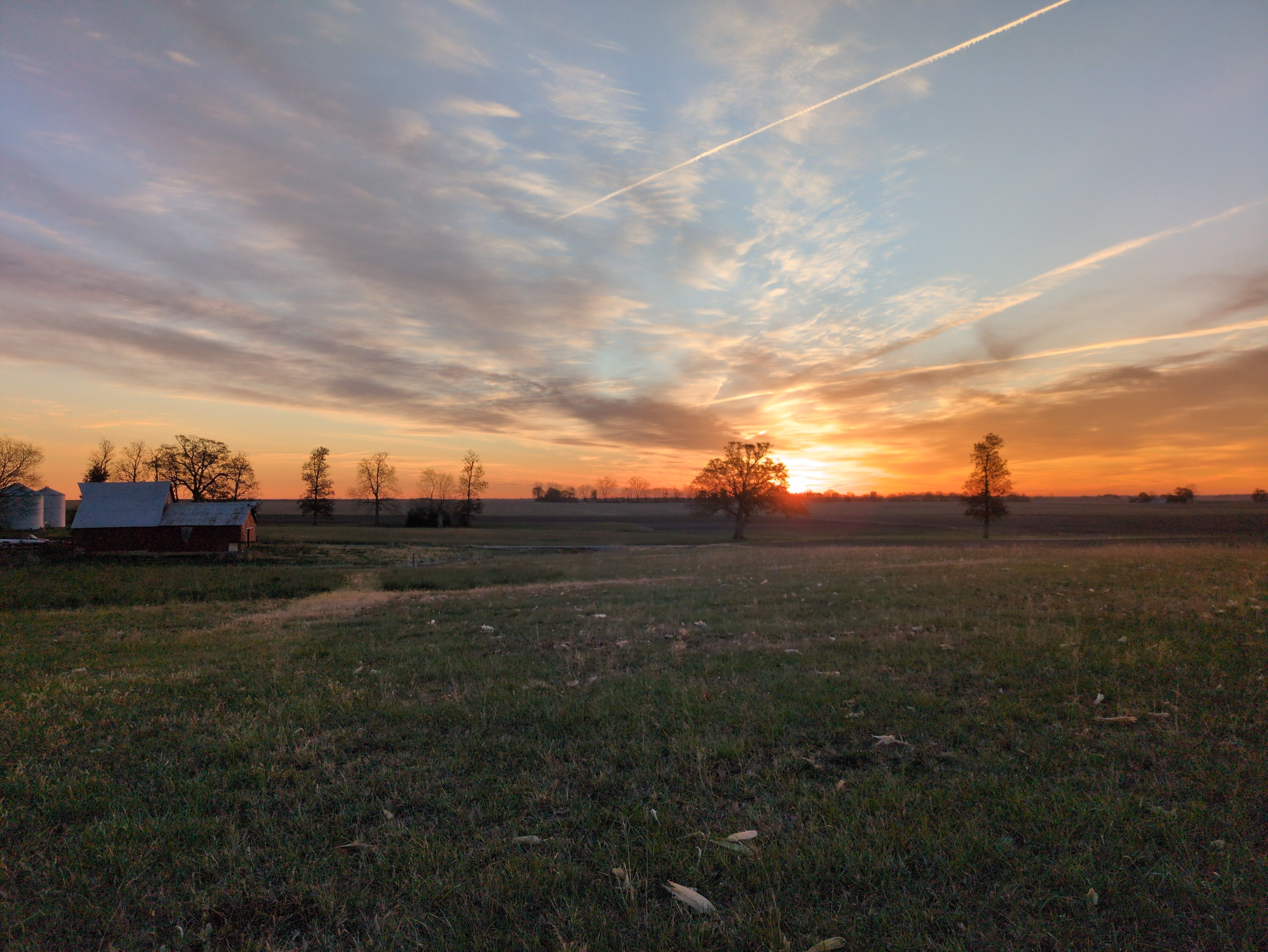 Sunrise over a mostly open field. The sun is just over the horizon, partially obscures by the sillhouette of a tree, tinging the lower half of the sky a range of mauves and oranges. A barn is visible in the distance.