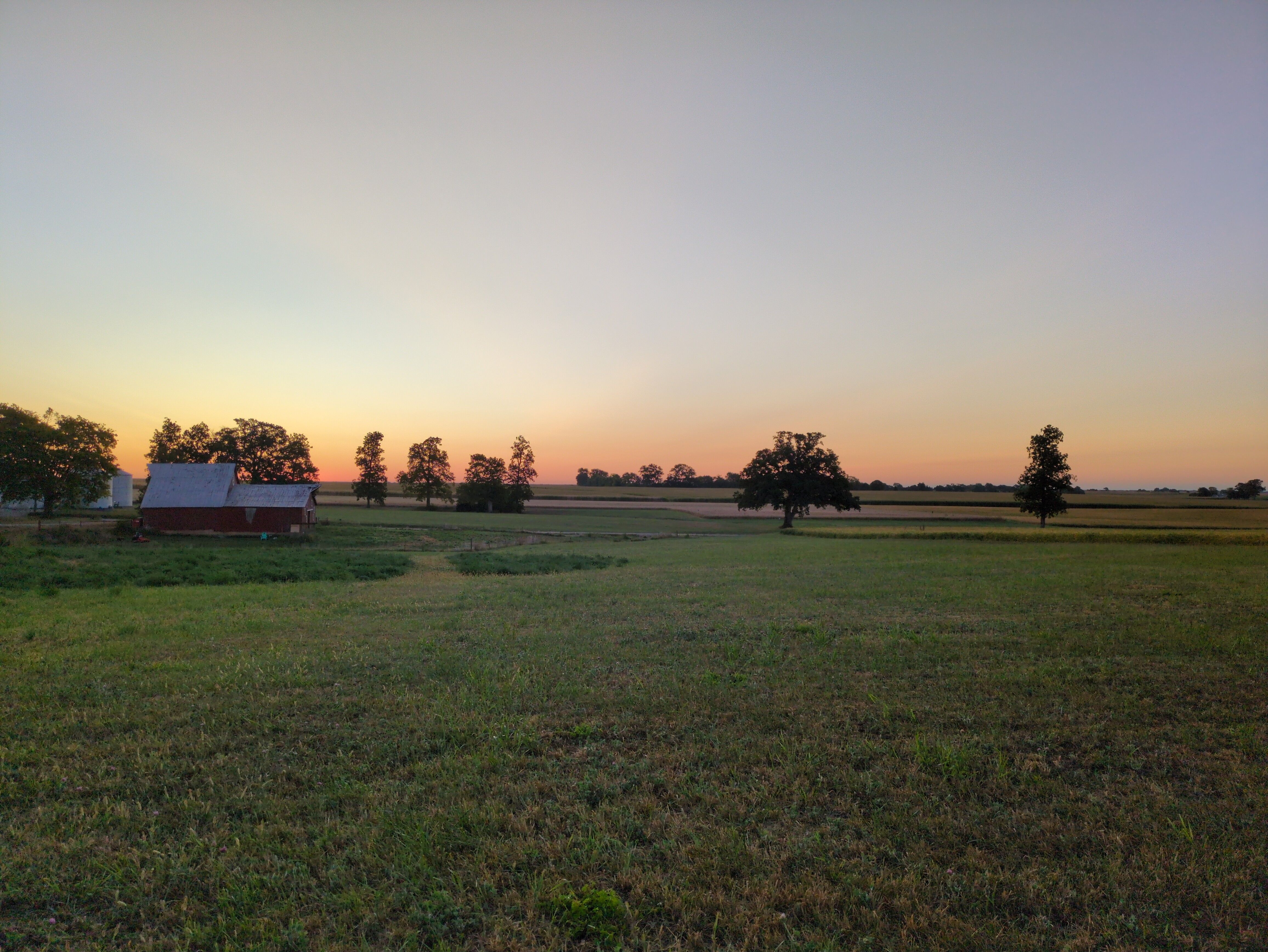 A photograph of an open field with a barn to the center-left of the frame. There are silhouettes of trees against a subtle, orange-peach sky.