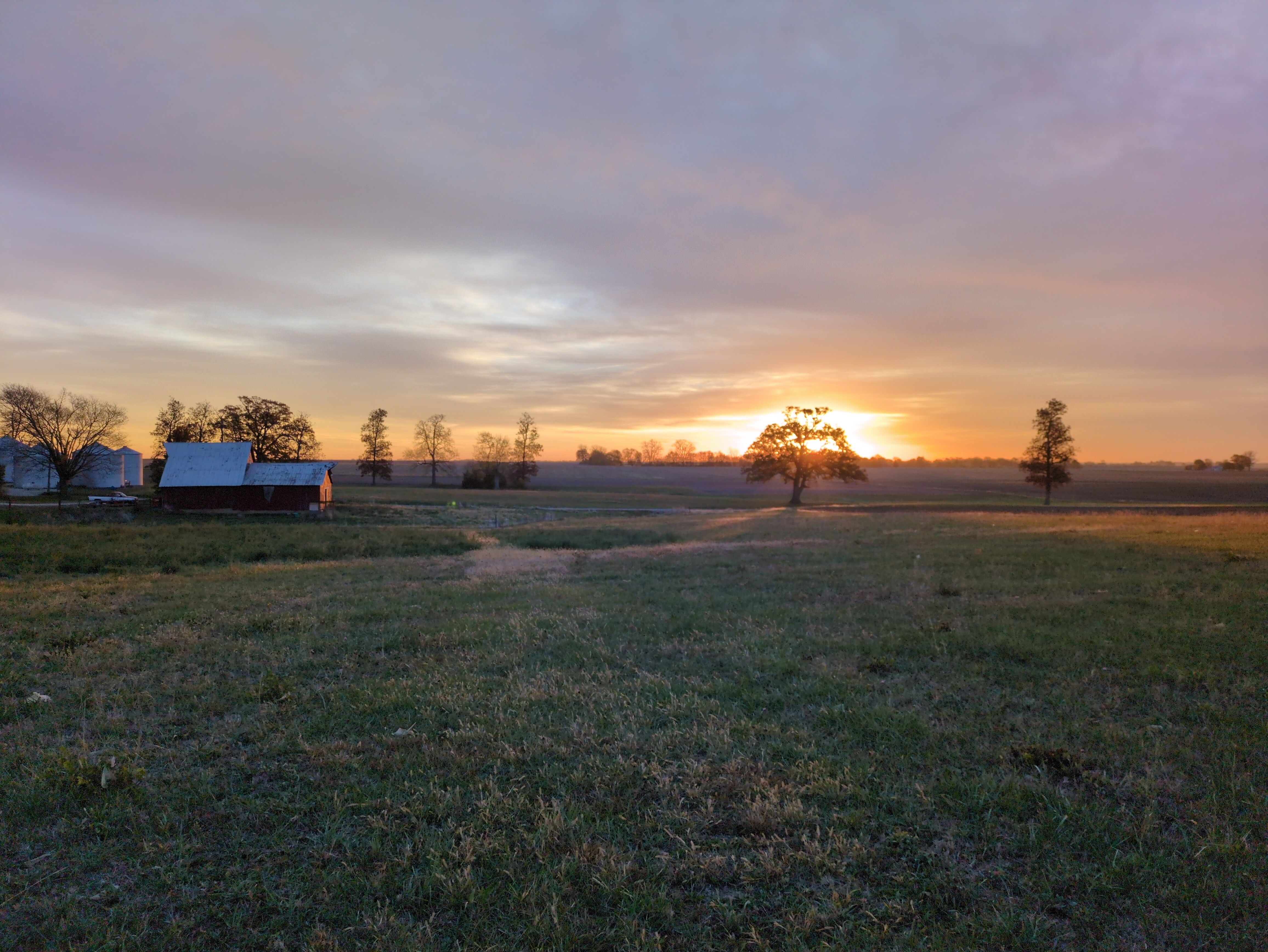Photo of an open field and big sky. Sky is hazy, but a sun is peeking through at the horizon, coloring the horizon in shades of deep orange. A barn and silhouttes of trees dot the horizon.