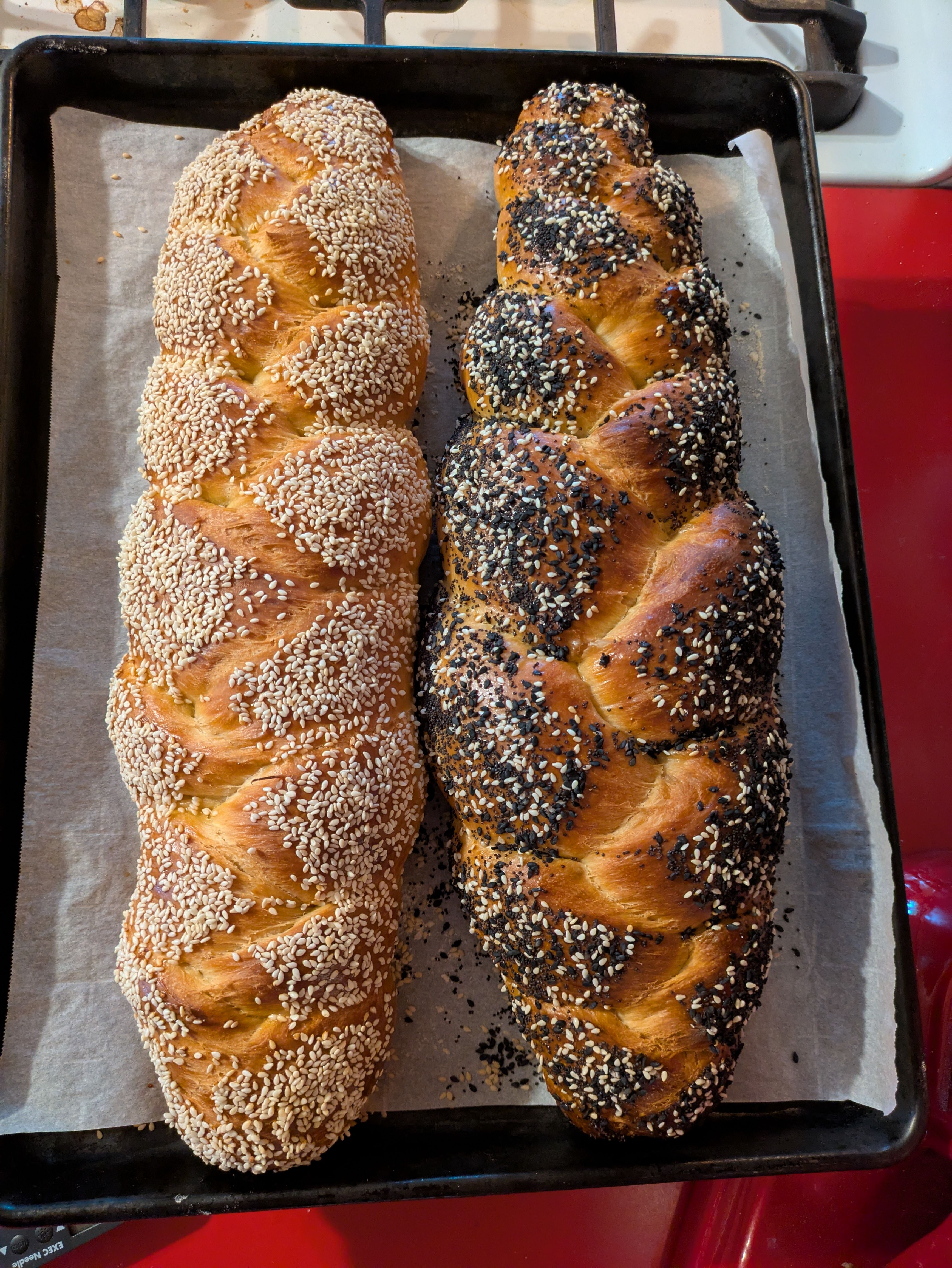 Photo of two long braided loaves of challah. One is covered in white sesame. The other is a mix of seeds. They are resting on a parchment-lined baking sheet.