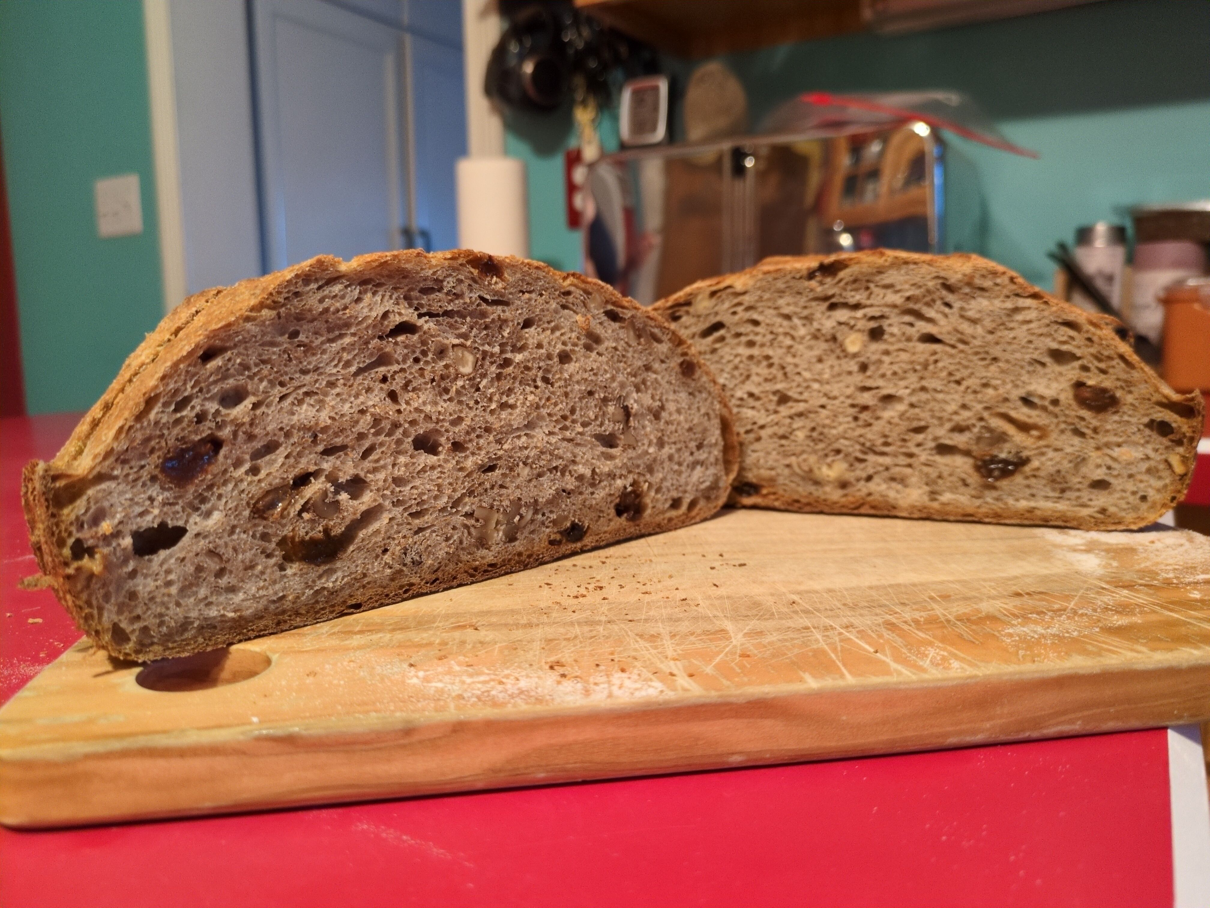 Two halves of a loaf of bread on a wooden cutting board, cut-side facing the camera. Raisins and walnuts are visible throughout the bread. The crumb is beige, the crust tan to golden.