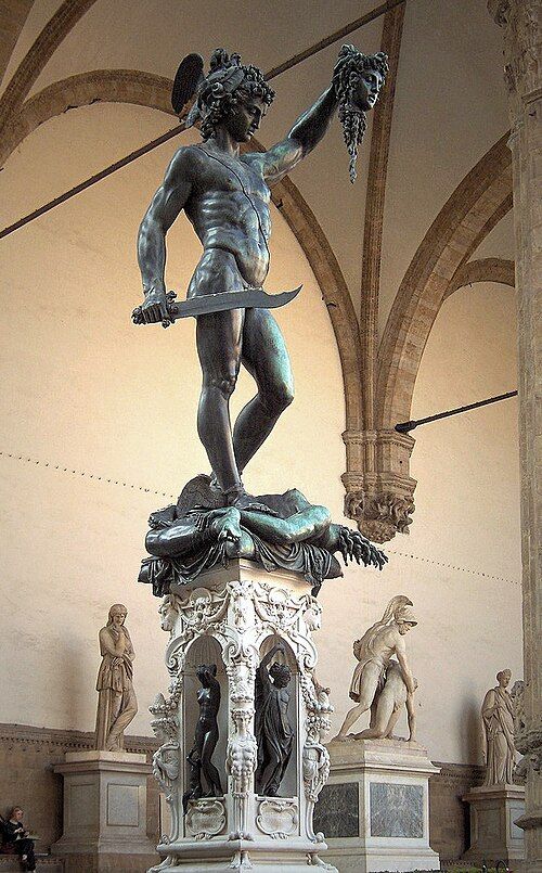 Photo of a sculpture of a muscular man holding up the head of a woman. His gaze is cast downward. He is stepping upon her body. In the background are other sculptures, as it appears to be in a museum.